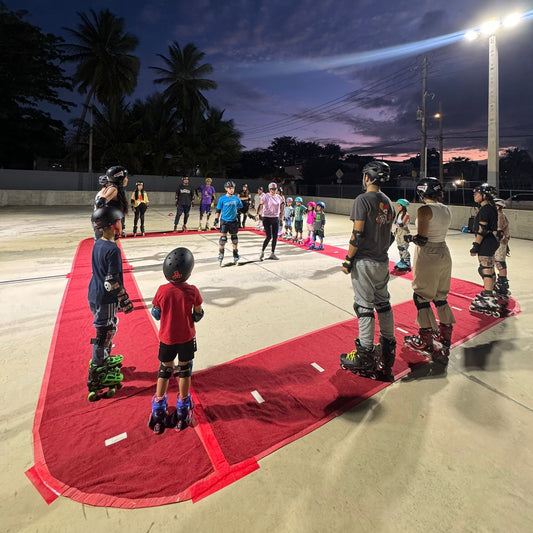 Group of people roller skating on a red track at night with palm trees in the background.